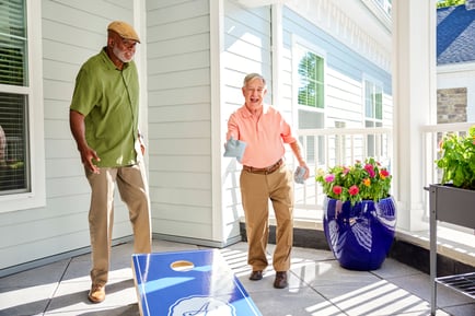 Two older adults laugh and play cornhole on a sunny outdoor patio surrounded by colorful flowers, showcasing the lively, social spirit of active senior living Herndon VA and senior living with dining and activities Herndon at Arbor Terrace Herndon.