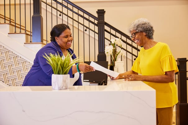 A smiling receptionist hands paperwork to a cheerful older woman at a front desk in an upscale lobby