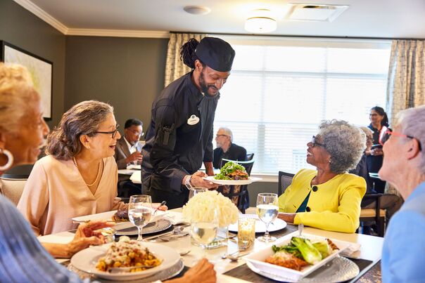A chef presents a fresh, beautifully plated dish to smiling residents at a sunlit table, highlighting the inviting dining experience at senior living in Citrus Park, FL.