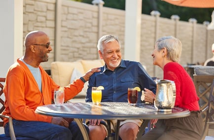 Three older adults smiling and laughing together at an outdoor patio table with glasses of iced tea, orange juice, and lemon water.