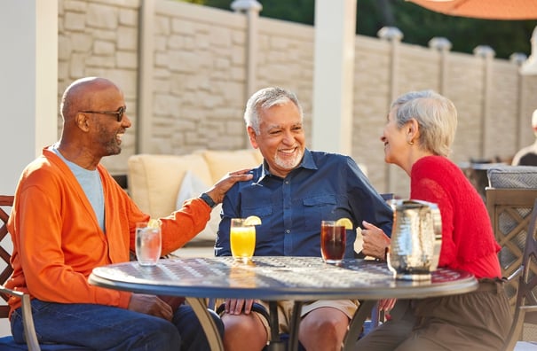 Three older adults smiling and laughing together at an outdoor patio table with glasses of iced tea, orange juice, and lemon water.