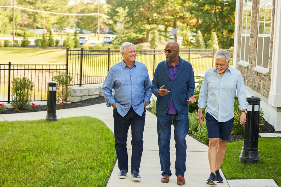 Three residents in casual button-up shirts walk and chat along a paved path lined with green lawn, red flowers, and bollard lights outside Arbor Terrace Highland Park. This reflects the active senior lifestyle and senior living with walking paths in Highland Park, IL.