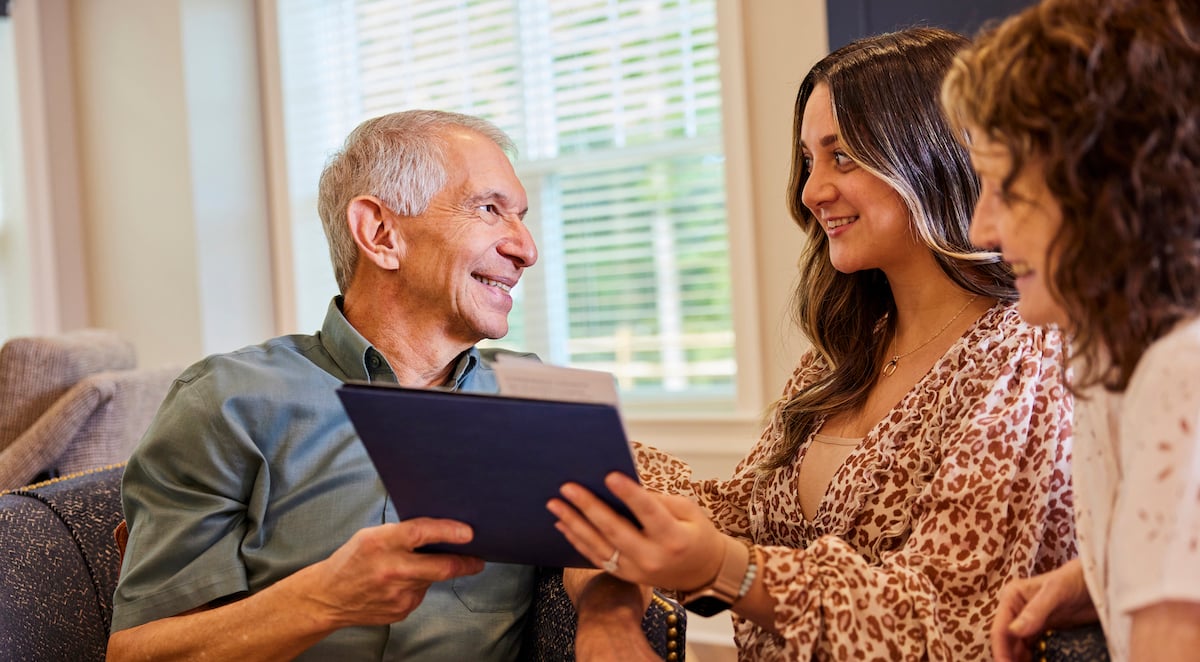 Staff members showing a resident a folder while resident sits in a gray chair. all are smiling