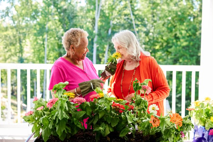 Two older women smile and tend to colorful blooms together on a sunny outdoor patio, enjoying one of the many senior-friendly activities near Fort Myers, FL, surrounded by lush greenery.