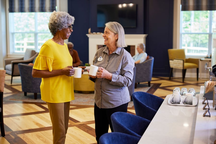 Two older women smiling and chatting while holding coffee mugs in a bright senior living community lounge, with a staff member wearing a name badge, cozy seating and a fireplace in the background.