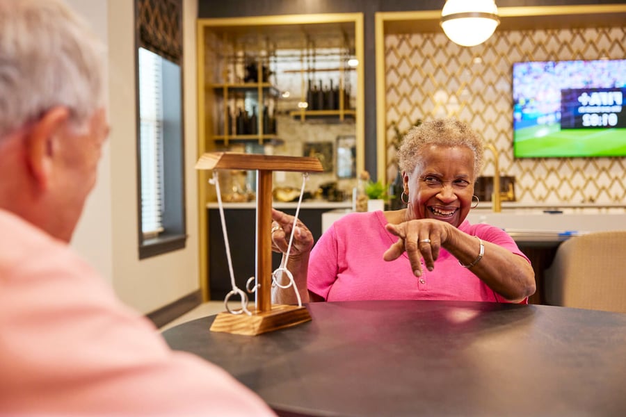 Two older adults share a warm, laughing conversation over a wooden puzzle game at a memory care community in Fort Myers, FL, seated at a table in a cozy, warmly lit lounge area.
