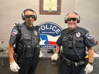 Two Lakeway Police officers wearing sensory simulation headsets and gloves smile for a photo in front of a Lakeway Police sign during a Dementia Live training session.