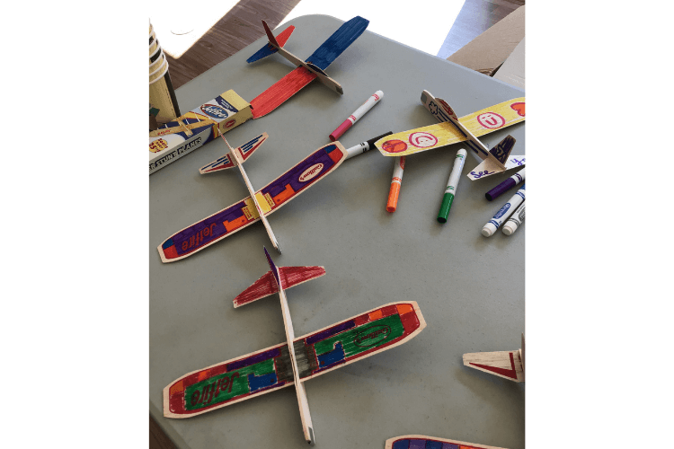 Colorful hand-decorated balsa wood glider planes spread across a table alongside markers — a creative activity celebrating National Aviation Day at senior living Lakeway Texas.