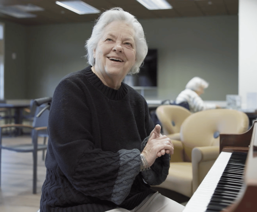 A resident smiles joyfully while seated near a piano in a spacious common area, reflecting the warm, dependable care and engaging senior lifestyle programs at this Acworth, GA community.