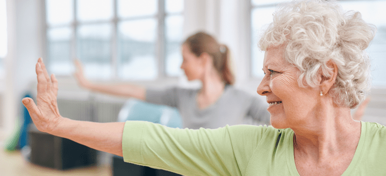 A smiling resident in a green top extends her arm during a stretching class at Arbor Terrace Highland Park, with an instructor mirroring the pose behind her. This reflects the active senior lifestyle and wellness-focused senior living in Highland Park, IL.