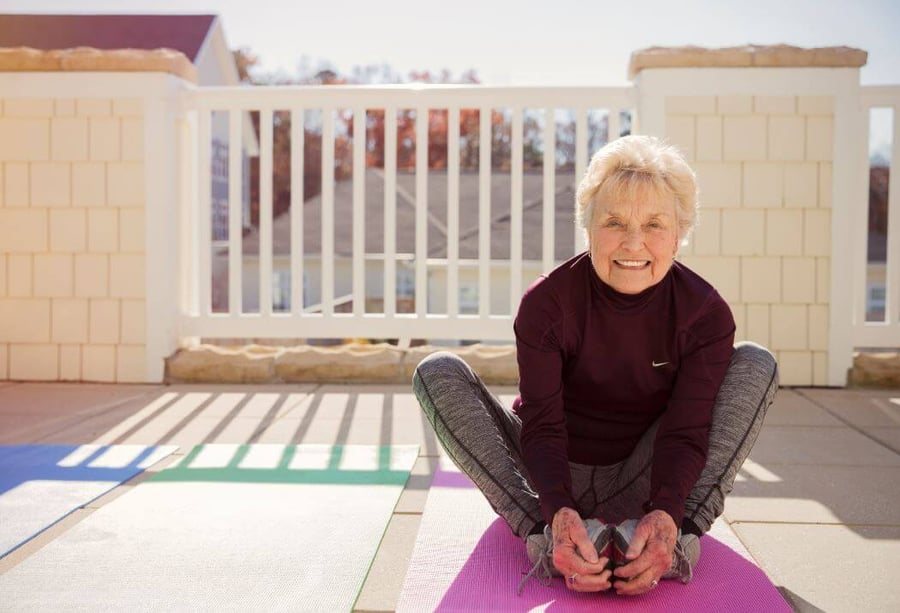 An older woman with short white hair sits on a purple yoga mat outdoors, smiling at the camera in a butterfly stretch pose. She is wearing a dark maroon Nike long-sleeve top and gray athletic leggings. Several colorful yoga mats are laid out on the patio around her, with a white fence and a rooftop visible in the background on a sunny day.
