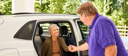 A staff member helps a smiling female resident out of a white vehicle surrounded by lush greenery, showcasing the reliable transportation services available at senior living Woodbridge VA.