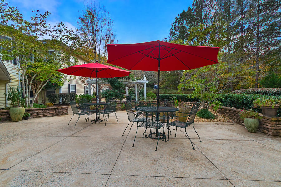A charming outdoor patio at Arbor Terrace Athens features wrought iron tables and chairs under bright red umbrellas, surrounded by mature trees and lush gardens at this top-rated senior living community in Athens, GA.