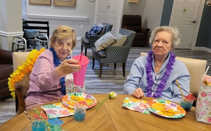 Two older adults wearing colorful leis enjoy a cheerful tropical-themed gathering at a memory care community in Athens, GA, with festive plates, bright drinks, and a warm common area visible in the background.