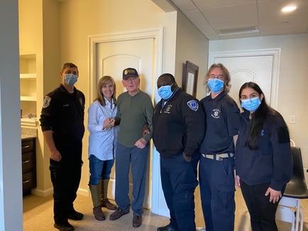 A senior resident is welcomed home to Arbor Terrace Mountainside by a group of smiling staff and first responders wearing masks, showcasing the caring community at this assisted living community in Mountainside, NJ.