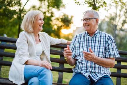 Two older adults sit on a sunny park bench, smiling and chatting together — reflecting the social and supportive memory care Lakeway TX offered at Arbor Terrace.