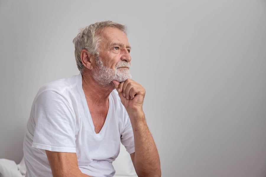 An older man with a gray beard rests his hand under his chin with a thoughtful expression against a neutral background, reflecting the focus on cognitive care and brain health at senior living communities in Athens, GA.