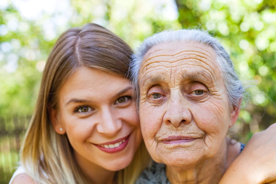 A younger woman and an older woman pose cheek to cheek outdoors, smiling amid lush greenery, reflecting the warm bonds at a senior living community in Suwanee, GA