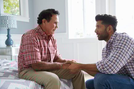 Two men sit face-to-face on a bed, holding hands in a heartfelt conversation in a softly lit bedroom, reflecting the compassionate family connections supported at assisted living Woodbridge VA.