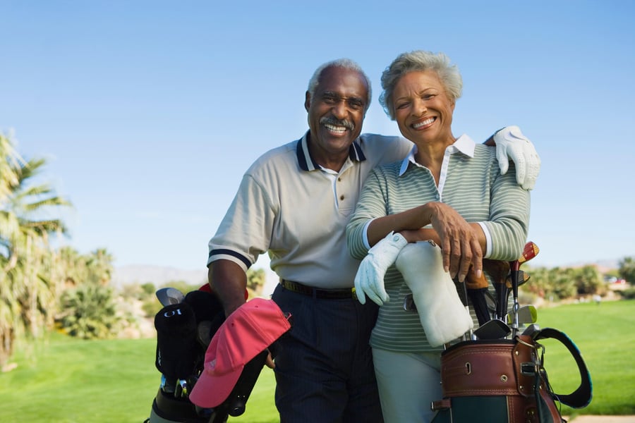 Two older adults smile warmly on a sunny golf course with their clubs, capturing the active, fulfilling lifestyle of independent living near golf courses Herndon and active senior living Herndon VA at Arbor Terrace Herndon.
