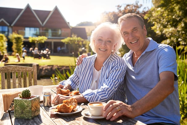 Two older adults smile and share a meal at an outdoor table surrounded by greenery and sunshine — person-centered memory care Lakeway TX at Arbor Terrace.