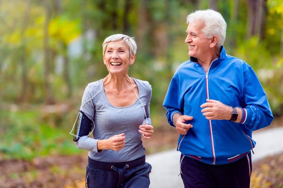 Two older adults jog side by side along a lush, tree-lined path, smiling and enjoying the outdoors together — embodying the vibrant, wellness-focused senior living Northern Virginia lifestyle at Arbor Terrace Herndon, a top independent living Herndon VA community
