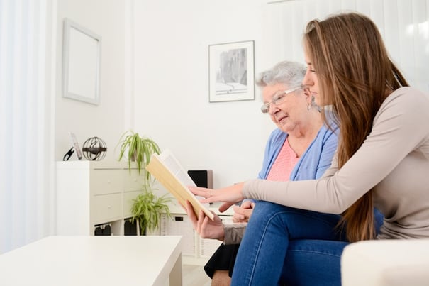 A younger woman and an older woman with glasses look together at an open book in a bright, plant-filled room, illustrating the supportive, personalized approach to memory care in Athens, GA.