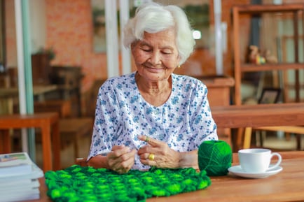 A woman smiles contentedly while crocheting at a wooden table with a cup of tea nearby, enjoying a peaceful activity at a senior living community in Suwanee, GA.