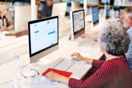 An older adult uses a desktop computer at a bright workstation alongside other seniors — staying connected through technology at senior living Lakeway Texas.