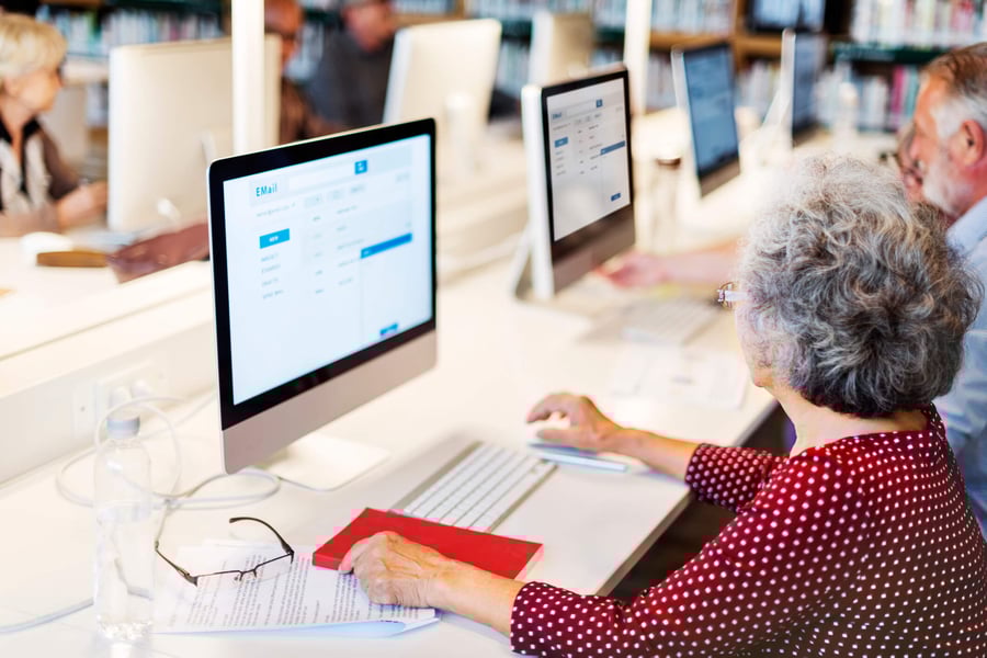 An older adult uses a desktop computer at a bright workstation alongside other seniors — staying connected through technology at senior living Lakeway Texas.