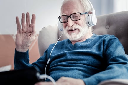 A cheerful older adult waves while enjoying music through headphones in a cozy chair, reflecting the active, connected senior lifestyle at Arbor Terrace Morris Plains, NJ.