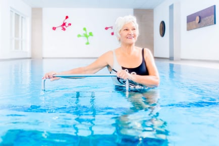 A smiling older woman stands in a bright indoor pool, gently gripping an exercise bar during aquatic therapy at a memory care community in Fort Myers, FL, with colorful wall decor in the background.