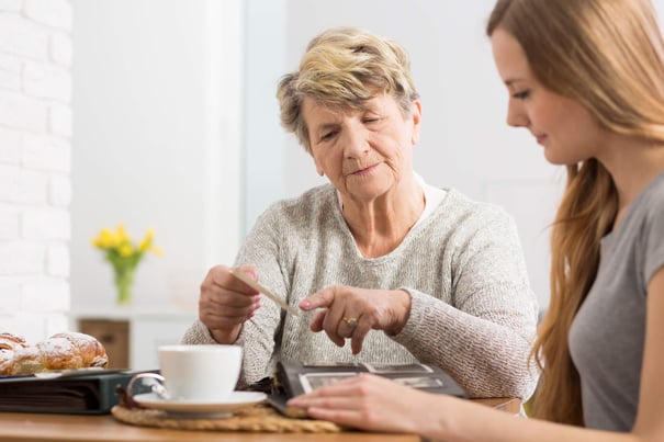 A younger woman sits beside an older woman at a table, gently assisting her as she stirs a cup of tea next to a pastry and yellow flowers, reflecting compassionate memory care in Fort Myers, FL.