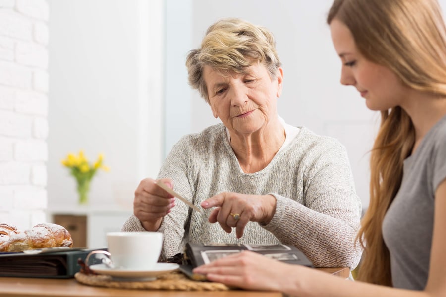 A younger woman sits beside an older woman at a table, gently assisting her as she stirs a cup of tea next to a pastry and yellow flowers, reflecting compassionate memory care in Fort Myers, FL.