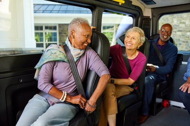 Three older adults smile and chat inside a community van, enjoying the senior living transportation services available in Fort Myers, FL, comfortably seated with seatbelts on.