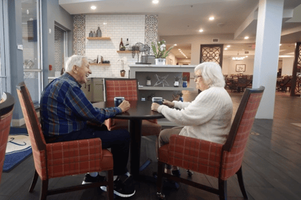 Two older adults share coffee and conversation at a small table in a warm, well-appointed dining area — the kind of connection found at an upscale assisted living community Lakeway TX.