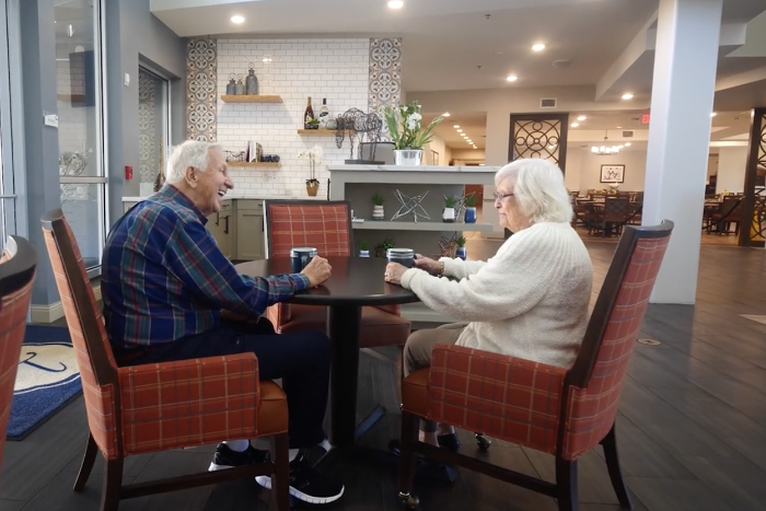 Two older adults share coffee and conversation at a small table in a warm, well-appointed dining area — the kind of connection found at an upscale assisted living community Lakeway TX.