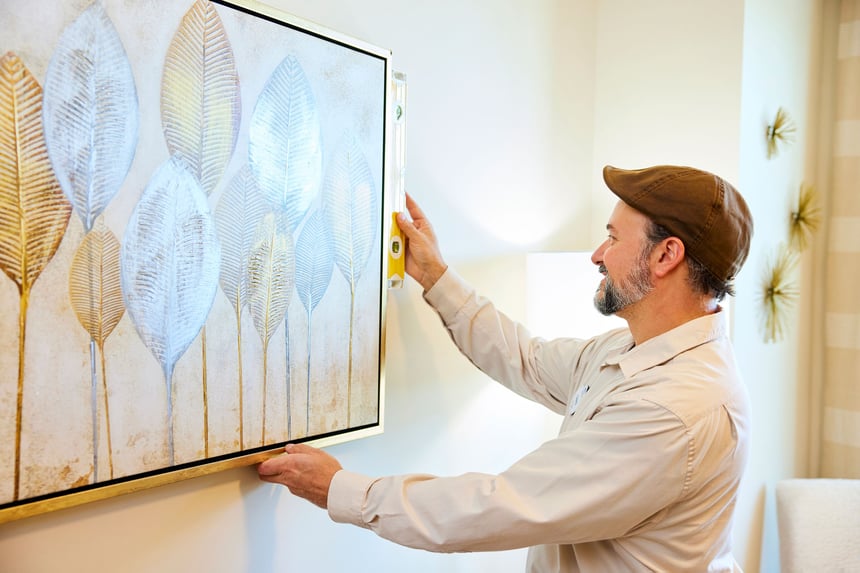 A maintenance staff member carefully levels a framed piece of artwork on the wall of a resident space at Arbor Terrace Athens, a senior living community in Athens, GA.