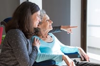 woman in grey shirt with her arm wrapped around a woman in a blue short points and looking out the window