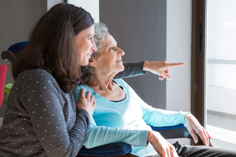 woman in grey shirt with her arm wrapped around a woman in a blue short points and looking out the window