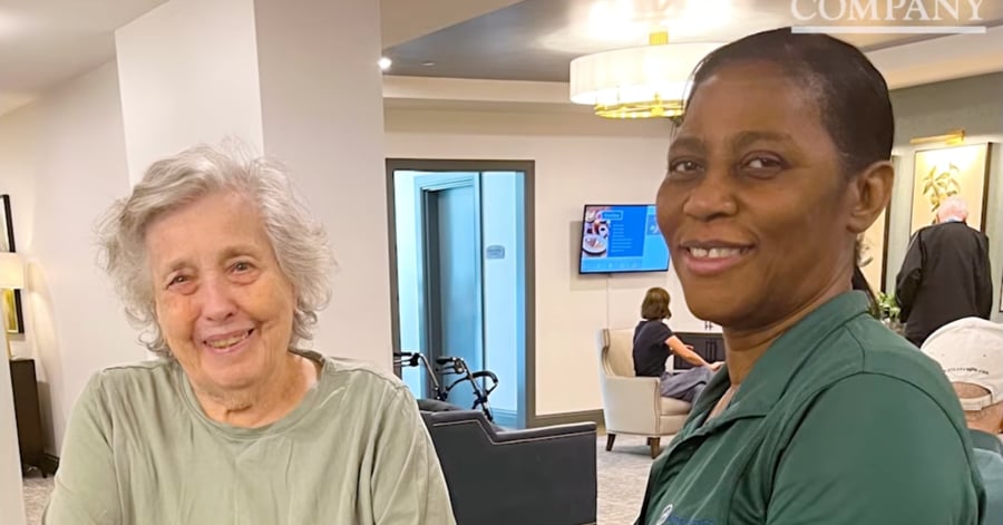 An elderly woman with white hair and a caregiver in a green polo shirt smile together in the common area of a senior living facility.