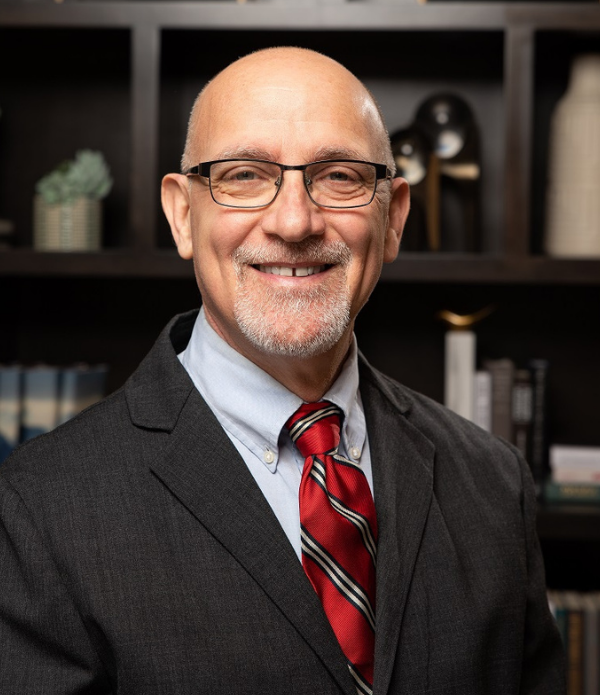 Christopher Dale, Executive Director at Arbor Terrace Highland Park, smiles warmly in glasses, a dark suit, and a red striped tie in front of bookshelves, leading a welcoming team for senior living Highland Park IL and personalized senior care Highland Park IL.