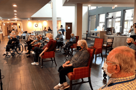 A group of seniors and staff wearing masks gathers in a warmly lit common room to enjoy a Classical Music Month event at Arbor Terrace Lakeway — vibrant senior care Lakeway TX.