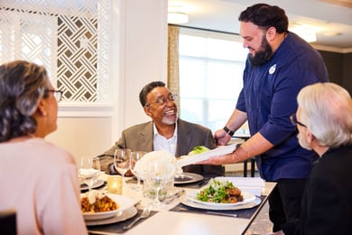 Staff member placing residents food on the table whole residents smile