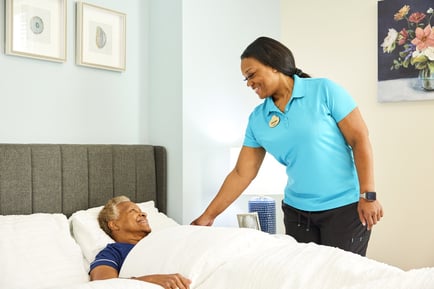 Woman assisting a male resident in bed with white sheets, grey headboard, and a picture on the wall