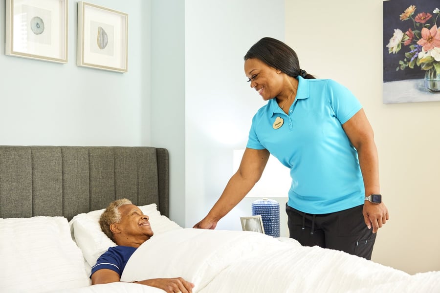 Woman assisting a male resident in bed with white sheets, grey headboard, and a picture on the wall