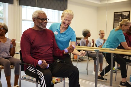 A staff member in a blue polo shirt smiles while assisting a seated older adult with hand weights during a fitness class at senior living in Fort Myers, FL, with other residents exercising in the background.