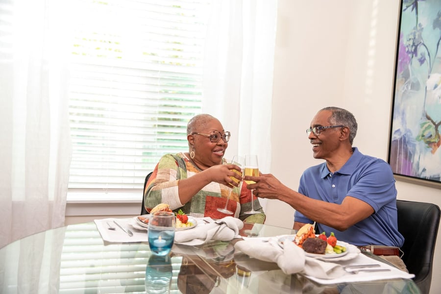 Two older adults share a joyful toast over a beautifully plated meal at a bright dining table, enjoying the social dining experience at an assisted living community in Athens, GA.
