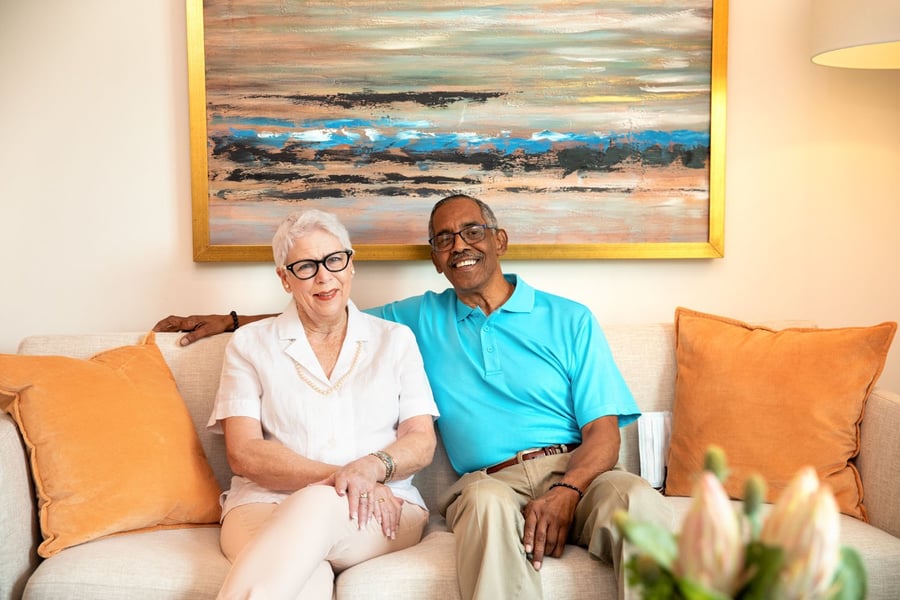 A couple sits together on a cozy sofa beneath a colorful painting, sharing a warm smile at a memory care community in Miami, FL where integrated care supports residents and their families with compassion.