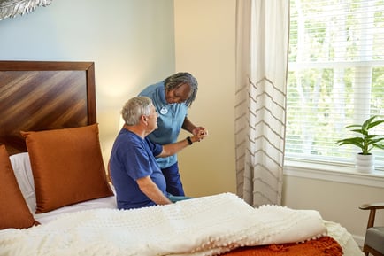 A care team member assists an older man seated on a bed in a warmly lit room, reflecting the attentive, personalized senior care provided at a Naperville, IL senior living community.